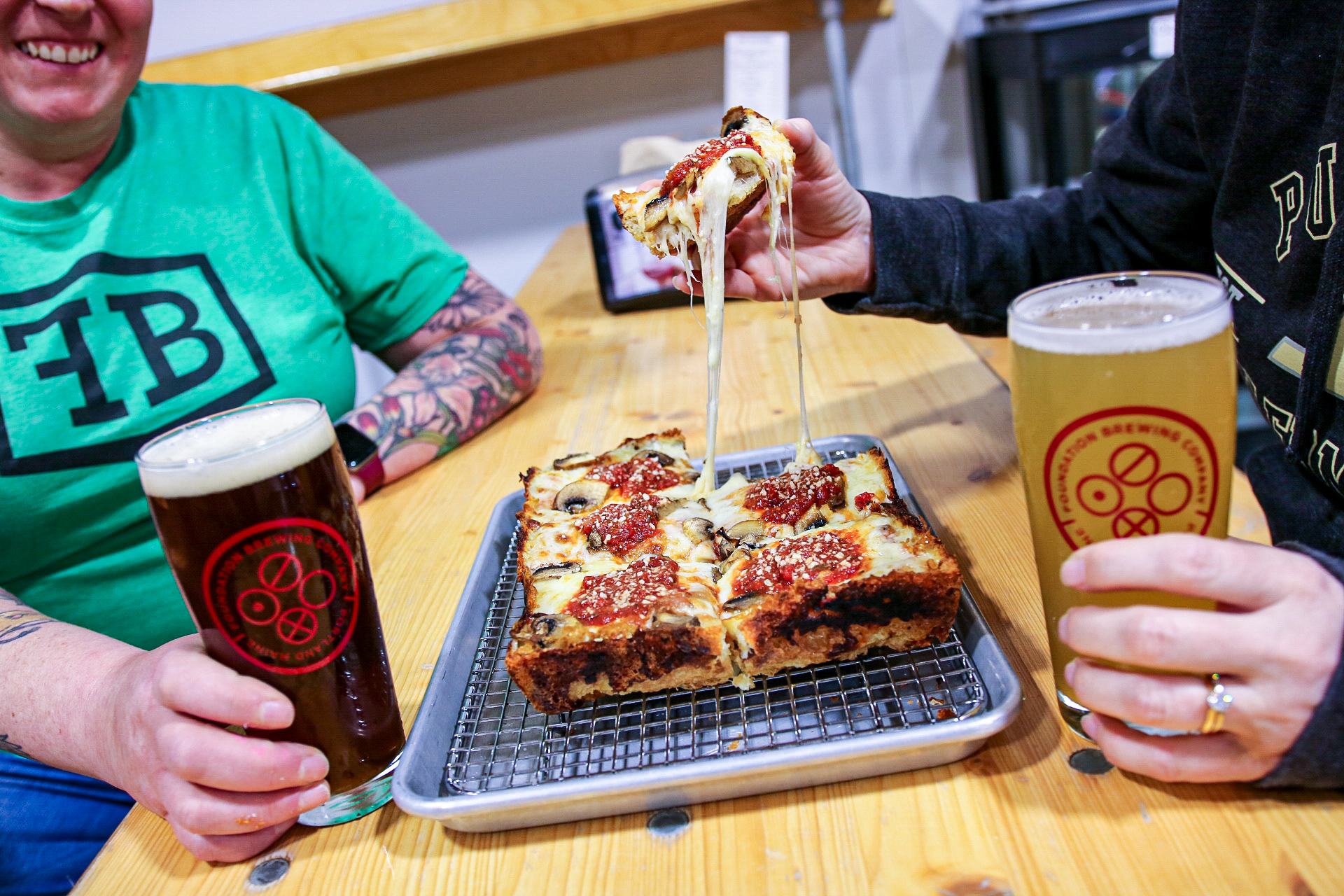 Two people sitting at a table. One is holding a beer with one hand and in the other is a slice of square pizza. The person across from them is holding a beer.